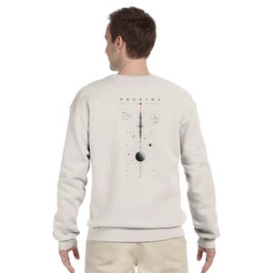 May include: A light beige long-sleeve sweatshirt featuring a graphic on the back. The design includes the text PROXIMA, a red star symbol, a vertical waveform, and a planetary system illustration with orbital rings and small circular celestial bodies.