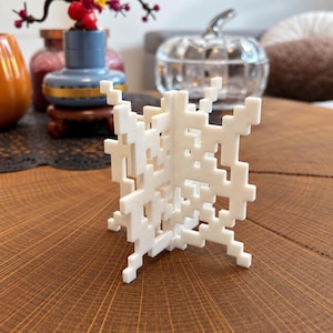 May include: A white, three-dimensional geometric sculpture featuring a pixelated, grid-like structure. The object is composed of interlocking, perpendicular panels that form a cross-shaped base and vertical planes with open, square-cut voids.