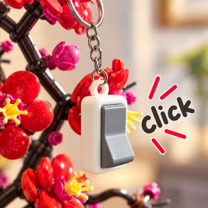 Pode incluir: Um porta-chaves com um pendente de interruptor de luz branco e cinzento, suspenso num anel de metal. O pendente está posicionado em frente a um ramo decorativo feito de blocos de construção de plástico vermelhos, rosas e amarelos com a palavra click.