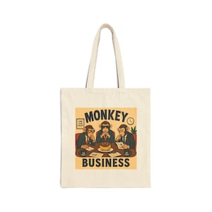 May include: A beige canvas tote bag featuring a graphic illustration of three monkeys in business suits sitting at a conference table. The text reads MONKEY BUSINESS, THINK PLAN EXECUTE, and Q1 GOALS. A banana sits on a plate in the centre.