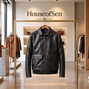May include: A black leather jacket with a front zipper, a classic collar, and two large patch pockets on the front, hanging on a wooden hanger from a gold metal rack in a retail store with the text HouseofSen displayed on the back wall.