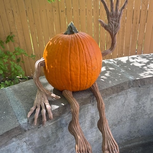 May include: A bright orange pumpkin with a green stem featuring attached brown, textured, branch-like limbs. The figure has two long arms with elongated fingers and two long legs with defined toes, posed in a seated position with one arm raised.