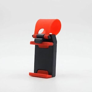 May include: A black and red mobile phone holder featuring a rectangular base with red silicone grips at the top and bottom, and a flexible red silicone loop at the top designed for attachment to a surface or object for hands-free viewing.