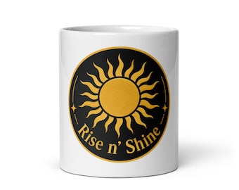 Puede incluir: Una taza de cerámica blanca con un emblema circular negro en el frente. El emblema muestra un sol amarillo dorado con rayos radiantes y el texto Rise n' Shine impreso en una fuente serif de color amarillo dorado a lo largo de la curva.