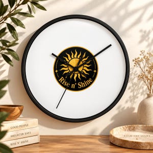 Puede incluir: Un reloj de pared redondo con marco negro y esfera blanca. En el centro hay un emblema circular negro con una ilustraci&oacute;n de un sol dorado y el texto "Rise n' Shine" en letras doradas. El reloj tiene tres manecillas negras y delgadas.