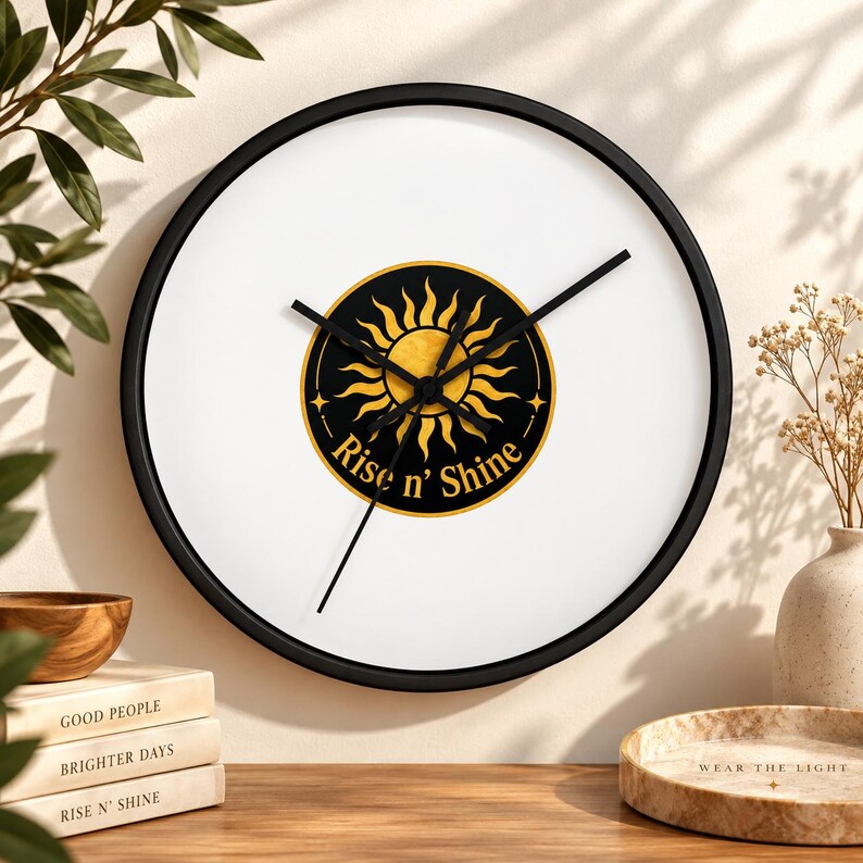 Puede incluir: Un reloj de pared redondo con marco negro y esfera blanca. En el centro hay un emblema circular negro con una ilustraci&oacute;n de un sol dorado y el texto "Rise n' Shine" en letras doradas. El reloj tiene tres manecillas negras y delgadas.
