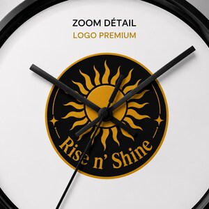 Puede incluir: Un reloj de pared redondo con marco negro y esfera blanca. El centro presenta un emblema circular negro con un gr&aacute;fico de sol dorado y el texto "Rise n' Shine" en letras doradas. Tres manecillas negras est&aacute;n situadas sobre el emblema central.