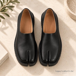 May include: A pair of black leather tabi shoes featuring a distinct split-toe design, a smooth upper surface, and a thick black rubber sole. The interior lining is a tan color. The text Legacy Leather Co Art is printed in the bottom right corner.