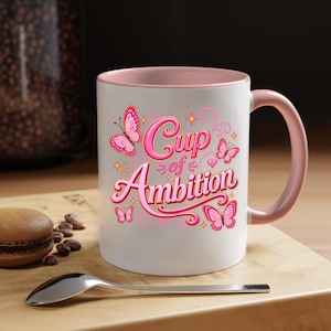 May include: A white ceramic mug with a pink interior and handle, featuring the text "Cup of Ambition" in pink script lettering surrounded by several pink butterflies, small stars, and decorative swirls printed on the front surface.