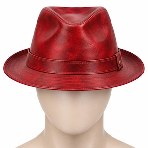 Puede incluir: Un sombrero fedora de color rojo intenso con una textura suave similar al cuero y un acabado ligeramente desgastado. El sombrero presenta una banda roja a juego alrededor de la copa, con un pequeño detalle de tachuela metálica y ala ancha.