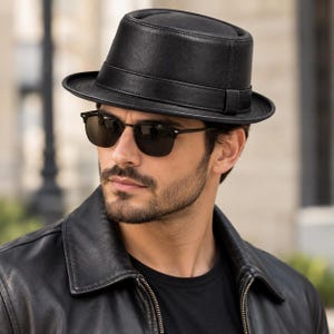 May include: A black leather pork pie hat with a flat crown and a narrow, upturned brim. The hat features a matching black leather band around the base of the crown. The material has a smooth, matte texture with visible stitching along the seams.