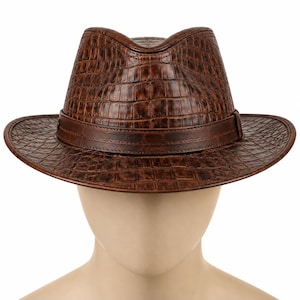 May include: A brown fedora hat featuring a textured crocodile skin pattern across the crown and brim. The hat includes a smooth, solid brown band encircling the base of the crown, providing a distinct contrast to the embossed reptilian texture.