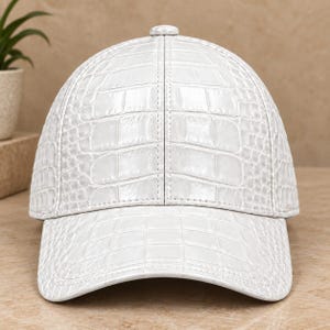 Puede incluir: Una gorra de béisbol blanca con un patrón texturizado de piel de cocodrilo en toda la superficie. La gorra tiene una corona estructurada con una costura vertical central, una visera curva y un pequeño botón a juego en la parte superior.