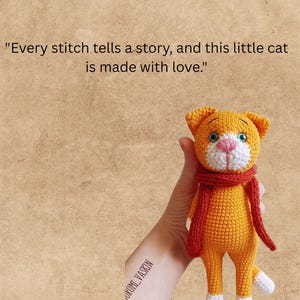 May include: A small orange crocheted cat toy with a white muzzle, white paws, and blue eyes, wearing a red scarf. The text above the cat reads: "Every stitch tells a story, and this little cat is made with love." The cat has a textured yarn finish.