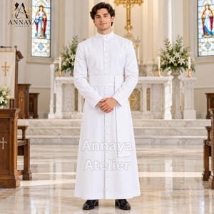 May include: A white cassock featuring a mandarin collar, long sleeves, and a full-length front button placket with matching white buttons. A wide, plain white fabric cincture is tied around the waist. The text Annaya Atelier is visible.