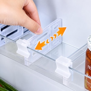 May include: A white plastic refrigerator shelf divider with a clip-on mechanism. The rectangular divider features a slotted design and is shown being adjusted horizontally along the clear plastic edge of a refrigerator door shelf.