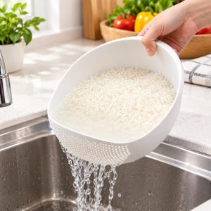 May include: A white plastic colander bowl with a perforated side panel, filled with white rice grains submerged in water. The water is actively draining through the small holes at the base of the bowl into a stainless steel kitchen sink below.