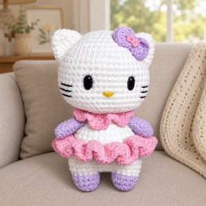 May include: A white crocheted plush toy featuring a large head with black oval eyes, a yellow nose, and three black whiskers on each side. It wears a purple bow, a pink ruffled collar, purple sleeves, a pink ruffled skirt, and purple feet.