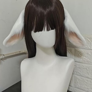 May include: A dark brown wig with straight hair and bangs, featuring large, plush white animal ears attached to the sides. The ears have a soft, light pink interior lining and are positioned symmetrically on the head of a white mannequin bust.