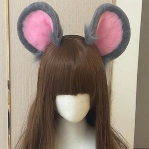 May include: A headband featuring two large, rounded mouse ears made of soft, plush material. The ears have a dark grey outer border and a bright pink inner section, attached to a brown wig on a mannequin head to display the accessory.