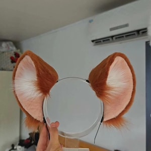 Può includere: Un paio di orecchie da animale in pelliccia sintetica attaccate a un sottile cerchietto nero. Le orecchie presentano uno strato esterno color arancio-marrone vibrante con fibre lunghe e morbide e una sezione interna rosa-bianco pallido.