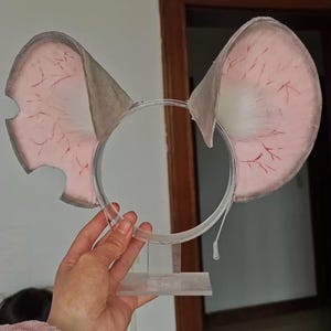 Puede incluir: Una diadema con dos orejas grandes y redondeadas con un interior rosa pálido detallado con finas venas rojas ramificadas. Los bordes exteriores de las orejas son de un gris apagado y la base de cada oreja tiene un mechón de pelo blanco.
