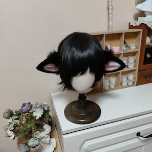 May include: A short black wig styled with straight bangs and attached animal ears. The ears are black on the exterior with soft pink inner detailing. The wig is displayed on a neutral-toned mannequin head mounted on a dark wooden pedestal base.