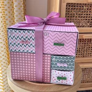 May include: A pink and green gift box with a purple ribbon. The box has four drawers with numbers 1, 2, 9, and 10 on them. The drawers have different patterns, including plaid, polka dots, and chevron.