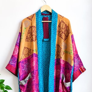 May include: A long-sleeved kimono-style robe featuring a patchwork design of vibrant magenta, mustard yellow, and deep maroon fabric panels. The garment includes a bright blue patterned trim along the front opening and two side pockets.