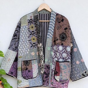 Op de afbeelding: Een patchwork jas met een verscheidenheid aan bloemmotieven en geometrische patronen in tinten paars, grijs en bruin. Het kledingstuk heeft lange mouwen, twee voorzakken met zwarte randen en een zwart-wit gestreepte voering bij de kraag.
