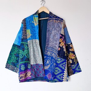 May include: A patchwork jacket featuring a variety of blue, green, and purple fabric panels with intricate floral, paisley, and geometric patterns. The garment is constructed with visible white kantha stitching throughout and hangs on a wooden hanger.