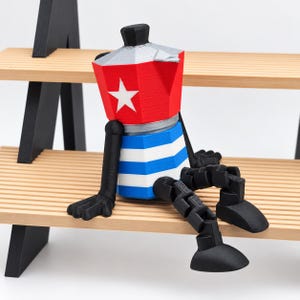 Peut inclure: Une figurine imprimée en 3D en forme de cafetière moka avec une partie supérieure rouge ornée d'une étoile blanche, une base rayée bleue et blanche, et des membres noirs articulés. L'objet possède un couvercle argenté et un bouton noir.