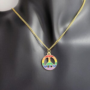 May include: Gold-tone necklace with a rainbow-coloured peace symbol pendant. The pendant is a circle with a peace sign inside, coloured red, orange, yellow, green, blue, and purple. The necklace is displayed on a black mannequin.