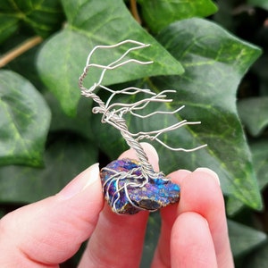 May include: A handmade wire tree sculpture with a silver trunk and branches, set on a colourful, iridescent stone. The tree is held against a backdrop of green leaves, creating a natural aesthetic.