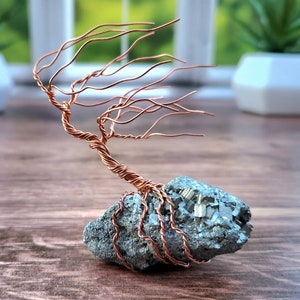 Op de afbeelding: Een koperdraad sculptuur van een boom met takken die in de wind waaien, geworteld in een grijze rots met pyrietkristallen.