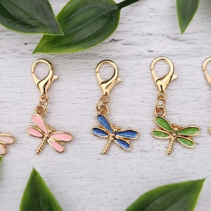 Peut inclure: Cinq breloques libellules avec des fermoirs dorés. Les libellules sont de différentes couleurs : rose clair, rose foncé, bleu, vert et noir. Les breloques sont présentées sur une surface en bois blanc avec des feuilles vertes.