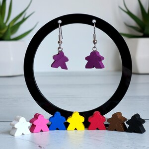 May include: A pair of purple game piece earrings suspended from silver hooks. The earrings are in the shape of a person. A black circular display stand holds the earrings. Below are colourful game pieces.