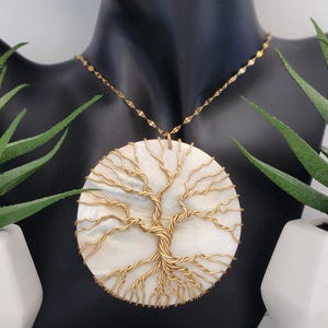 May include: A gold-coloured necklace featuring a circular pendant with a tree of life design. The pendant has a white, pearlescent background with a gold wire tree overlay. The necklace is displayed on a black mannequin.