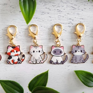 May include: Six cat-themed charms with gold-coloured clasps. Each charm features a different cartoon cat design in various colours, including pink, calico, grey, and black. Green leaves are scattered around the charms.
