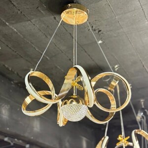 May include: A gold and white pendant light fixture featuring multiple curved, ribbon-like rings suspended from a circular gold ceiling canopy. A central spherical light element with a textured, faceted surface hangs in the middle of the rings.