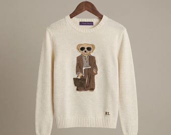 Op de afbeelding: Een crèmekleurige trui met lange mouwen en ronde hals, voorzien van een centrale afbeelding van een teddybeer met een zonnebril, een bruin pak en een klein tasje. De beer is geborduurd. Onderaan staan de letters RL in bruin geborduurd.