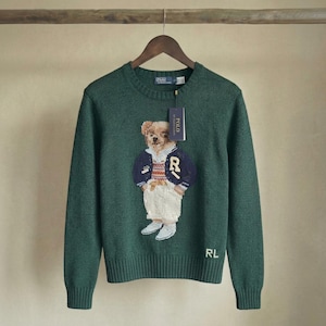 May include: A dark green long-sleeve crewneck sweater featuring a central graphic of a teddy bear wearing a navy blue varsity jacket, a patterned sweater, and white pants. The text POLO is printed on the attached tag and RL is on the hem.