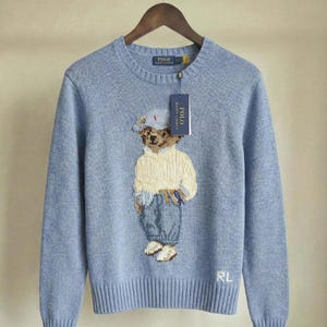 Op de afbeelding: Een lichtblauwe trui met lange mouwen en ronde hals, voorzien van een gebreide afbeelding van een teddybeer in een cr&egrave;mekleurige trui, blauwe broek en een pet. De trui heeft een Polo Ralph Lauren label en de letters RL in wit geborduurd.