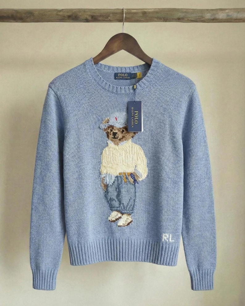 Op de afbeelding: Een lichtblauwe trui met lange mouwen en ronde hals, voorzien van een gebreide afbeelding van een teddybeer in een cr&egrave;mekleurige trui, blauwe broek en een pet. De trui heeft een Polo Ralph Lauren label en de letters RL in wit geborduurd.