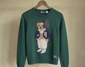 Op de afbeelding: Een donkergroene trui met lange mouwen en ronde hals, voorzien van een gebreide afbeelding van een teddybeer in een marineblauw vest, een gestreept overhemd en een witte broek. De beer heeft een R op de borst. De letters RL zijn wit geborduurd.