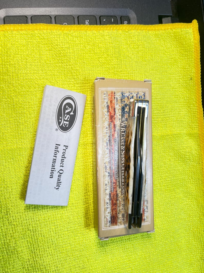 May include: A folding pocket knife with a textured cream and brown handle, resting on its rectangular cardboard box labeled W.R. Case & Sons Cutlery Co. Next to it is a white paper insert with the Case XX logo and Product Quality Information text.