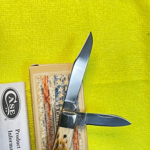 May include: A folding pocket knife with two silver-colored steel blades and a textured brown and cream handle. The knife is positioned on a tan box featuring an American flag design and the text W.R. CASE, alongside a white paper insert labeled CASE XX.
