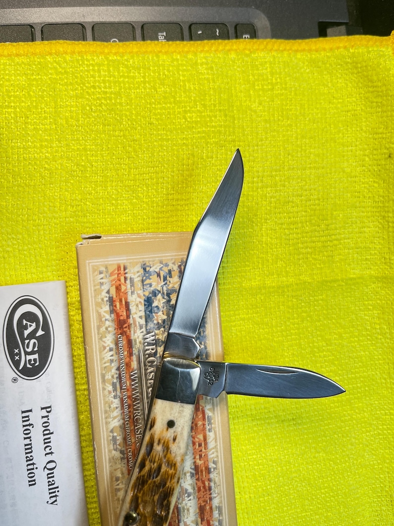 May include: A folding pocket knife with two silver-colored steel blades and a textured brown and cream handle. The knife is positioned on a tan box featuring an American flag design and the text W.R. CASE, alongside a white paper insert labeled CASE XX.