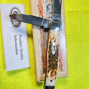 May include: A folding pocket knife with two silver blades and a textured brown and cream handle featuring a metal inlay. The knife rests on a rectangular box with a flag pattern and text, accompanied by a white paper labeled Product Quality Information.