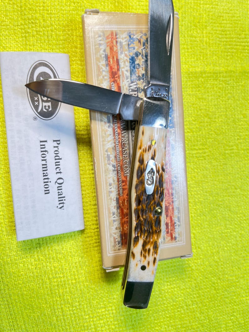 May include: A folding pocket knife with two silver blades and a textured brown and cream handle featuring a metal inlay. The knife rests on a rectangular box with a flag pattern and text, accompanied by a white paper labeled Product Quality Information.